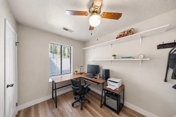 Room featuring wood-finish flooring, a window with horizontal blinds, a white door, a ceiling fan with light, and two white wall-mounted shelves