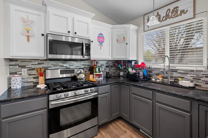 Corner kitchen with granite-look countertops, stainless steel appliances, mosaic tile backsplash, white upper cabinetry, and contrasting gray lower cabinetry