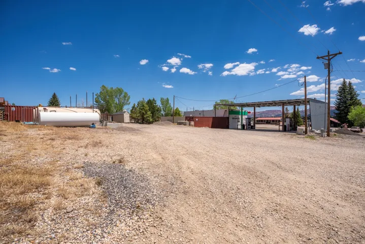 Gravel lot with utility infrastructure, featuring a large white storage tank, multiple small outbuildings, and a covered fueling station with visible pumps