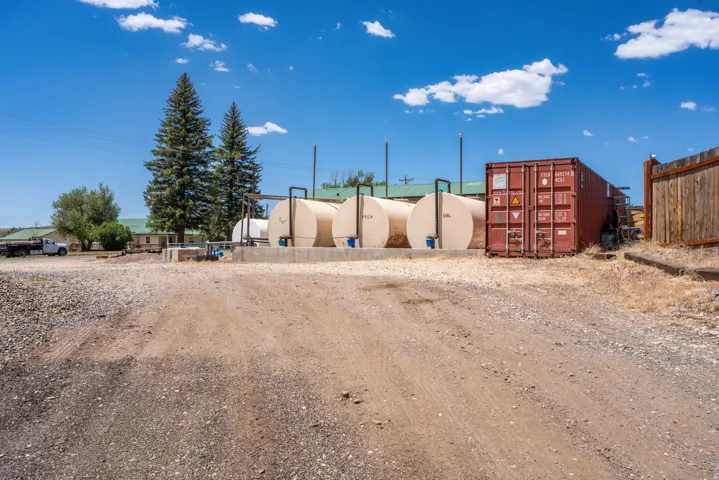 Industrial-grade storage area featuring three large cylindrical tanks, an adjacent shipping container, a gravel ground surface, and a wood fence boundary