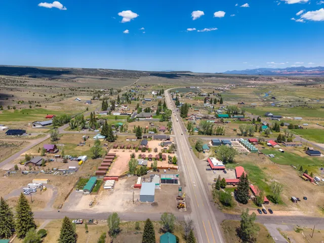 Expansive rural landscape featuring a central highway, scattered residential structures, mature trees, and distant mountainous terrain under a clear blue sky