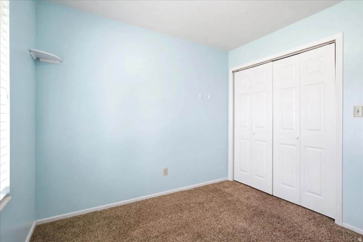 Bedroom featuring light blue walls, brown carpet flooring, white bi-fold closet doors, white baseboards, and a white window frame with blinds