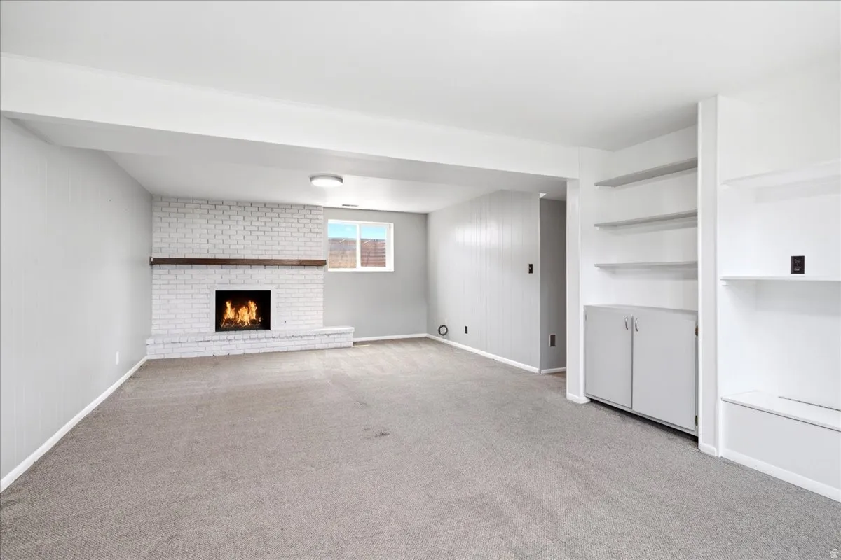 Spacious room featuring a white-painted brick fireplace with a wood mantle, neutral carpet flooring, and built-in shelving with cabinetry