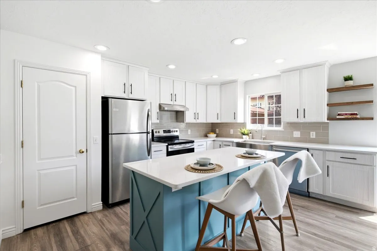 Bright kitchen featuring white cabinetry, stainless steel appliances, a center island with a white countertop and blue base, wood-finish flooring, and a subway tile backsplash