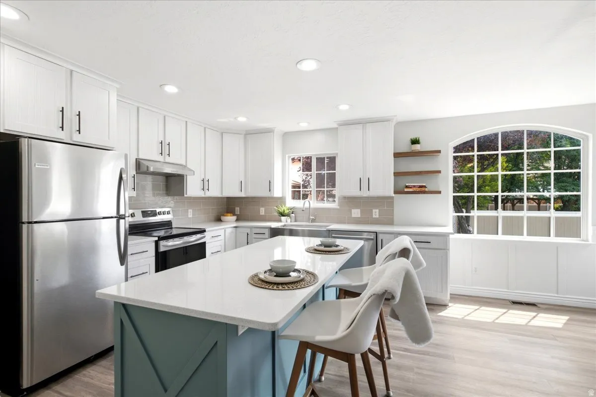 Modern kitchen featuring white cabinetry, stainless steel appliances, a center island with a light-toned countertop, wood-finish flooring, and a large arched window with grid detailing