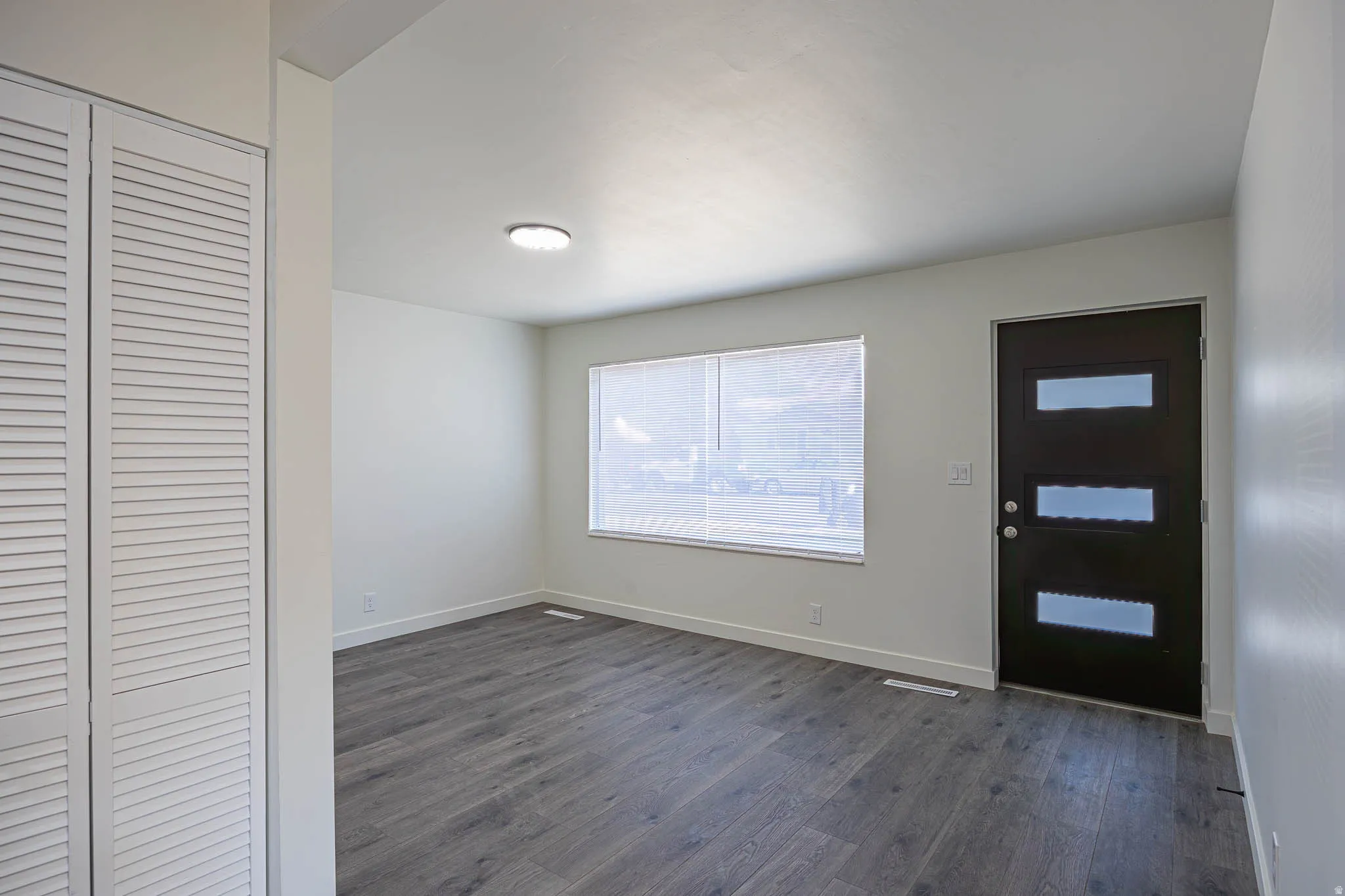 Entry area featuring wood-finish flooring, a contemporary dark entry door with frosted glass inserts, a window with horizontal blinds, a flush-mount ceiling light fixture, and white louvered bifold closet doors