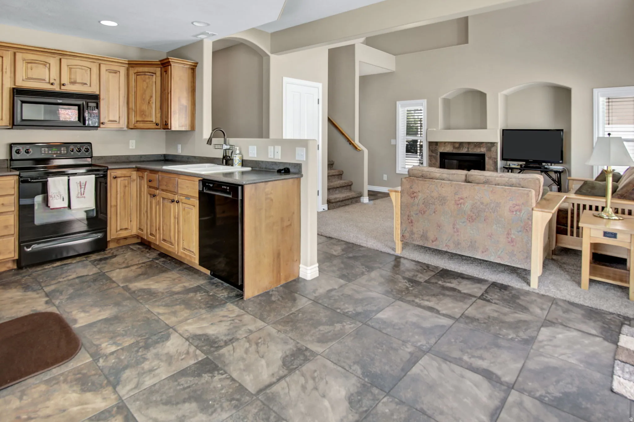 Open-concept kitchen featuring wood cabinetry, black appliances, a double basin sink, and stone-finish tile flooring