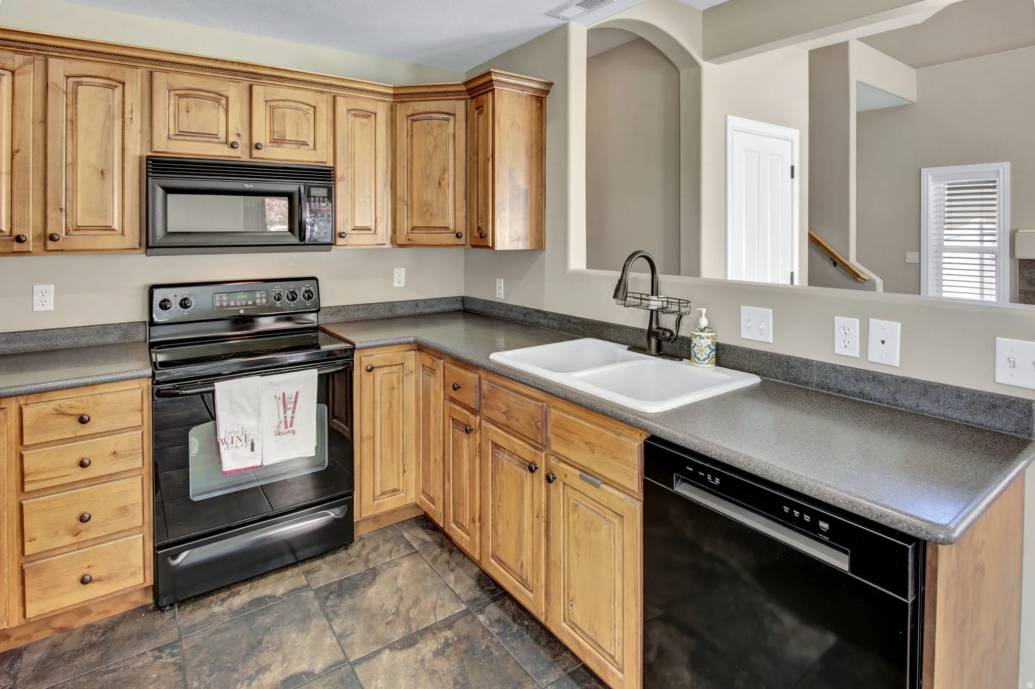 Kitchen featuring wood cabinetry, dark countertops, black appliances, a double basin sink with a gooseneck faucet, and tile flooring