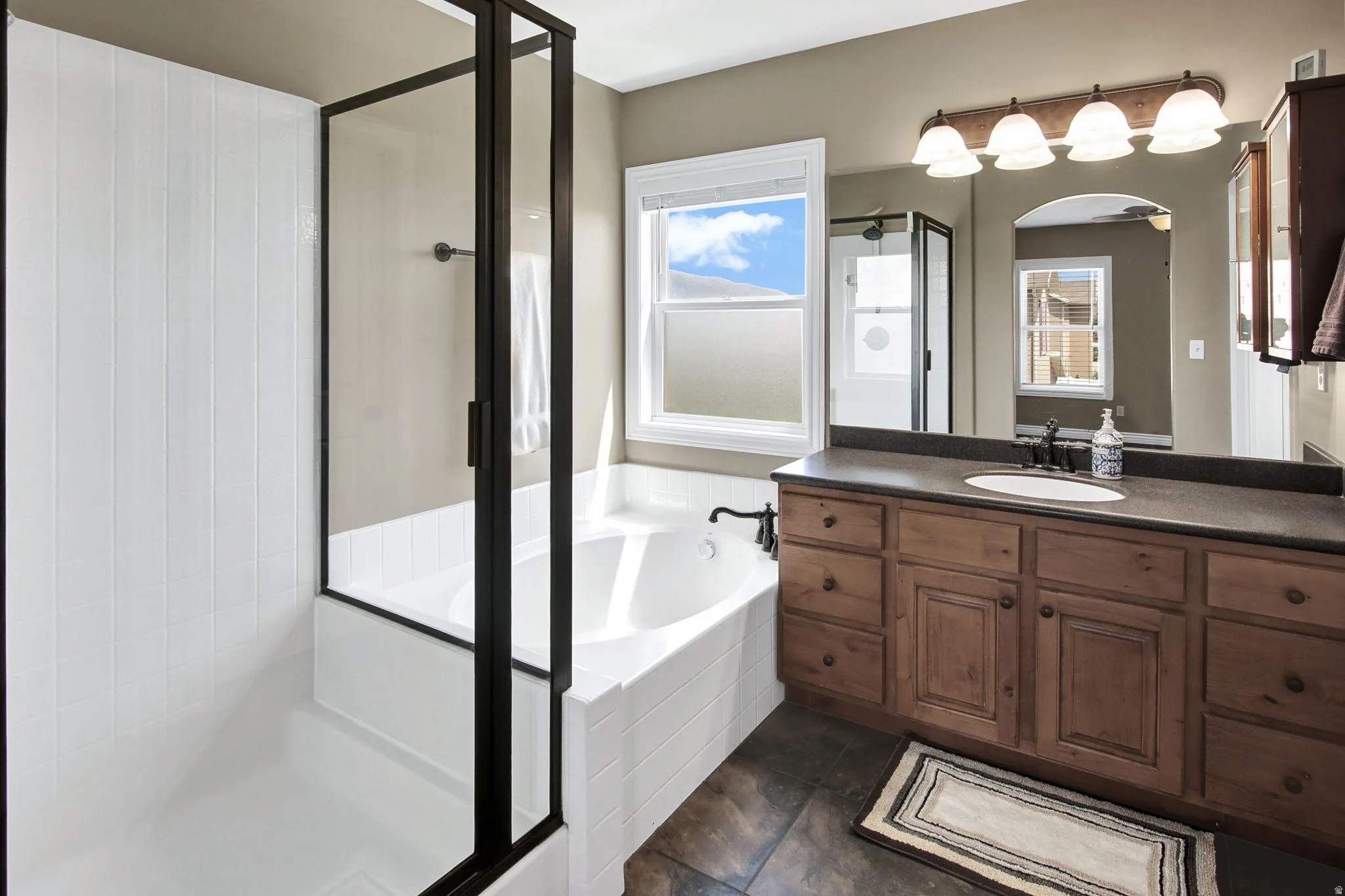 Spacious bathroom featuring a deep soaking tub with white subway tile surround, a separate shower with a black-framed glass enclosure, and a wood-finish vanity with dark countertop