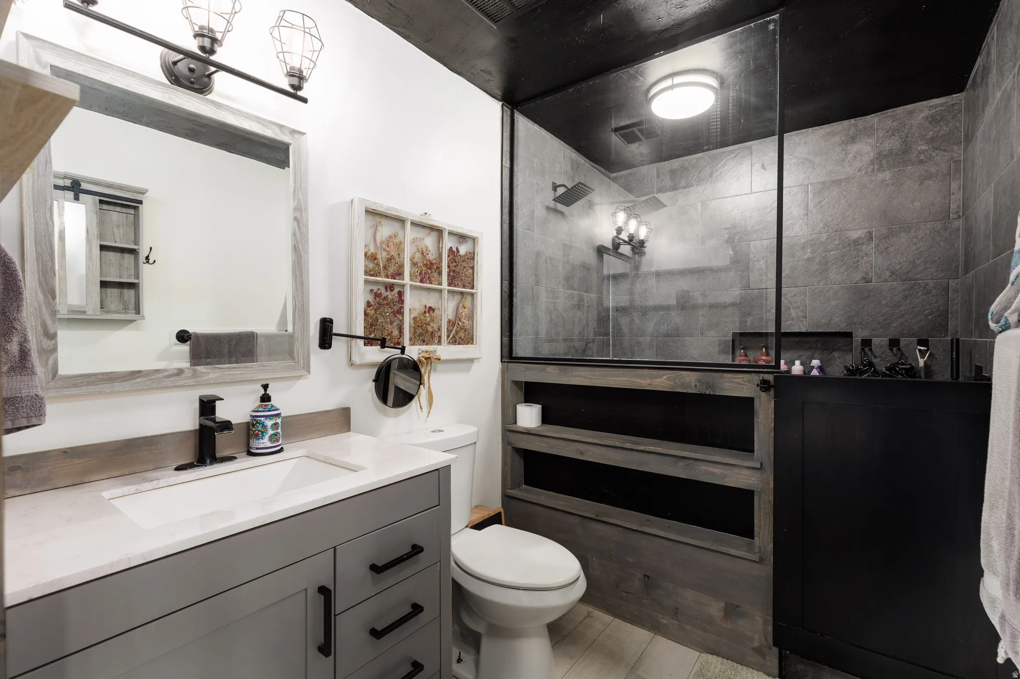 Modern bathroom featuring a glass-enclosed shower with dark stone-look tile walls, a vanity with a white countertop and gray cabinetry, light wood-finish flooring, and a contemporary black ceiling