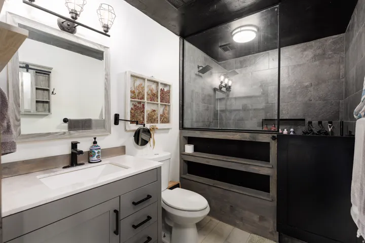 Modern bathroom featuring a glass-enclosed shower with dark stone-look tile walls, a vanity with a white countertop and gray cabinetry, light wood-finish flooring, and a contemporary black ceiling