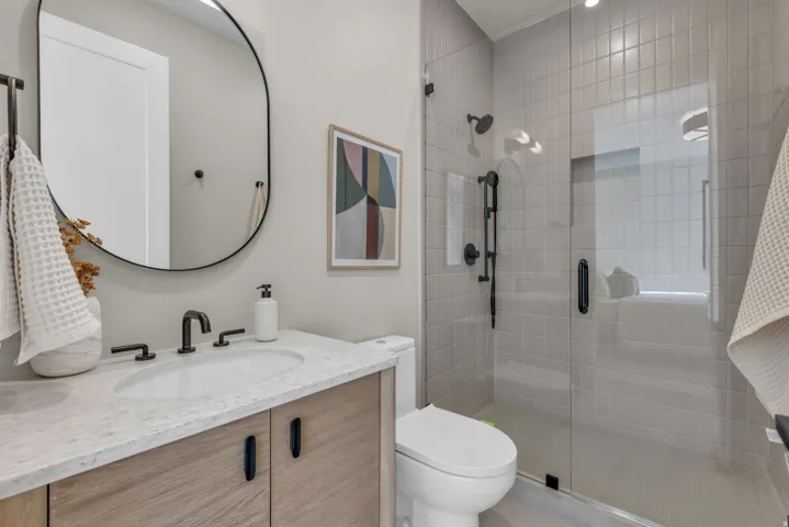 Bathroom featuring a wood-finish vanity with a light-toned countertop, an oval-shaped mirror, and a glass-enclosed shower with subway tile surround and matte black fixtures