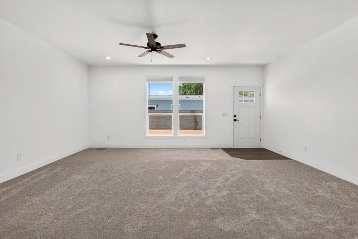 Spacious room featuring neutral carpeting, white walls, a multi-pane front door, a large window, and a ceiling fan with integrated lighting