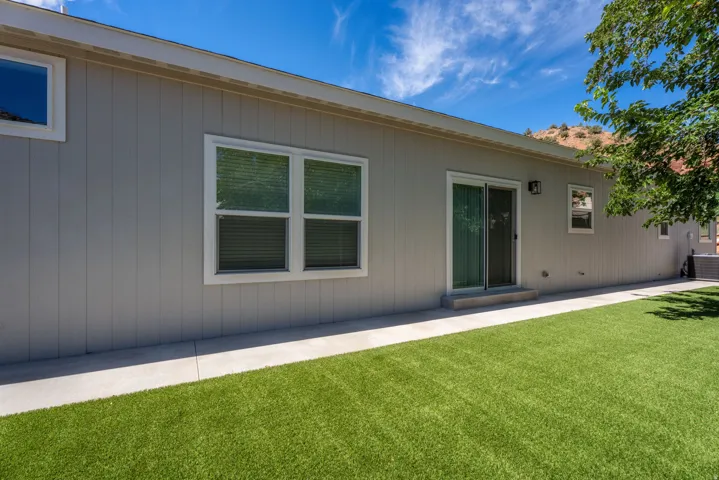 Gray vertical panel siding with white trim, multiple windows with blinds, sliding glass door, concrete walkway, and artificial turf lawn