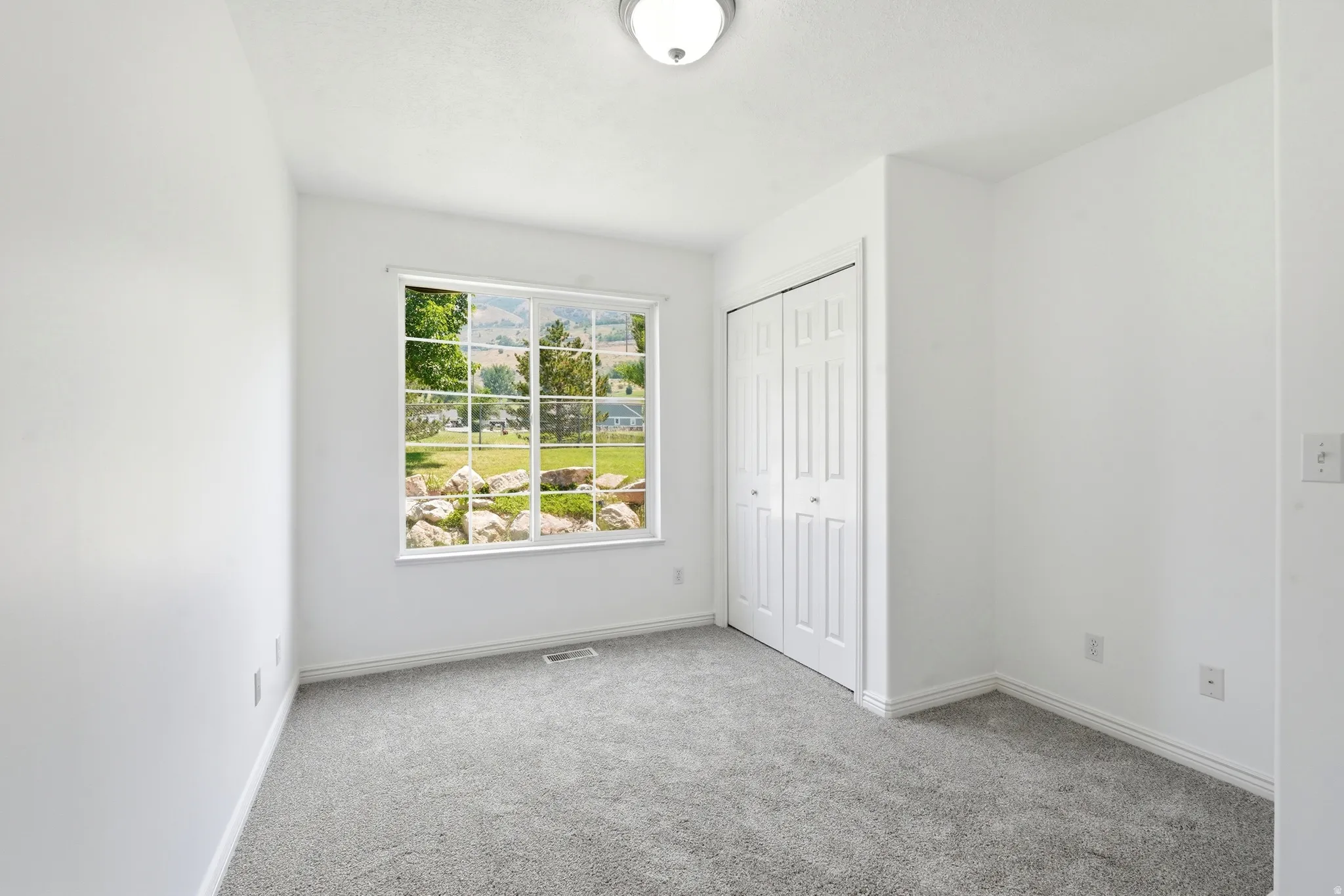 Light-filled room featuring a large window overlooking a landscaped exterior, plush gray carpeting, white walls, a ceiling-mounted light fixture, and a spacious bi-fold door closet