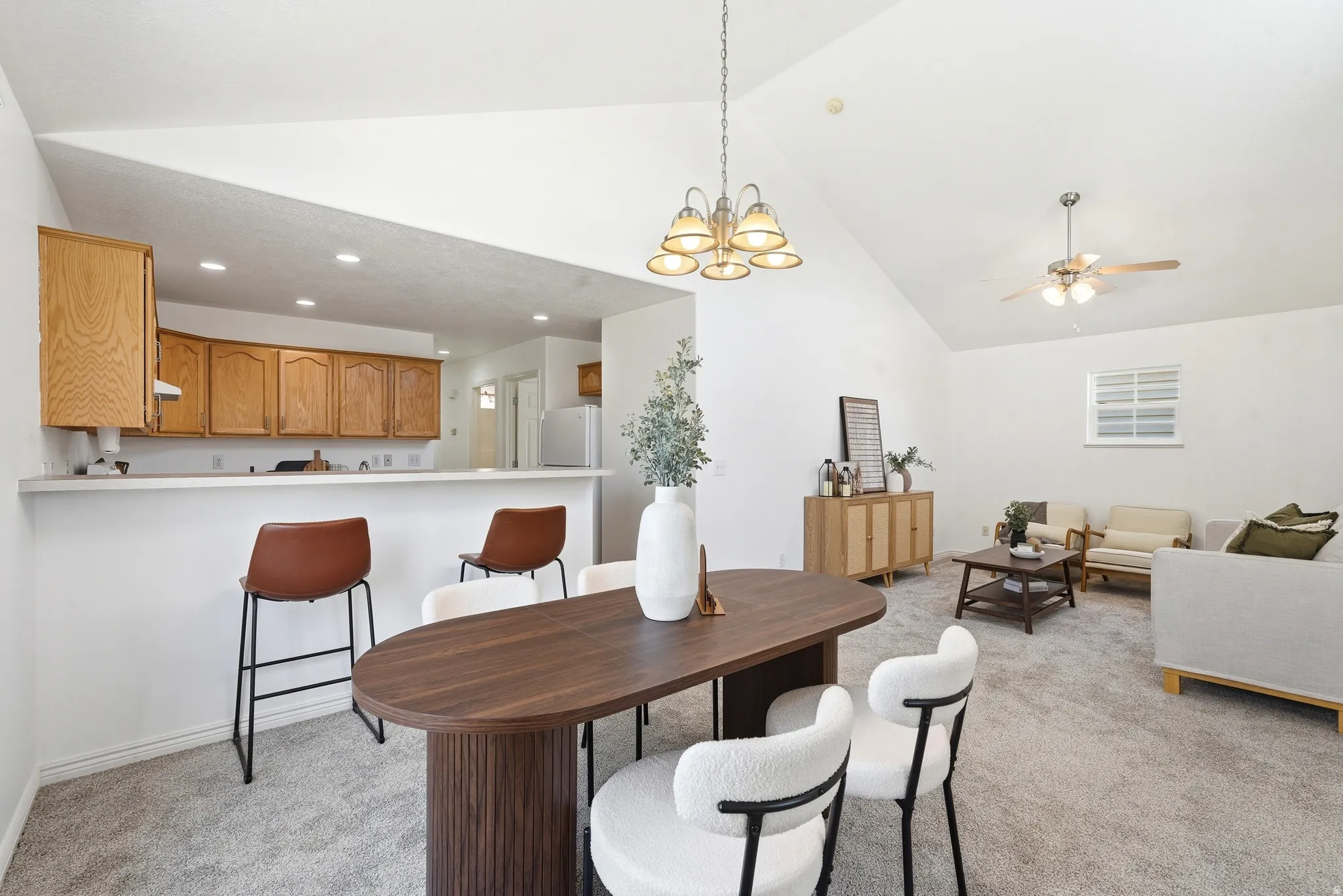 Vaulted ceilings enhance this open-concept room featuring a dining area with a modern chandelier, a kitchen with wood cabinetry and a breakfast bar, and a carpeted living space with a ceiling fan