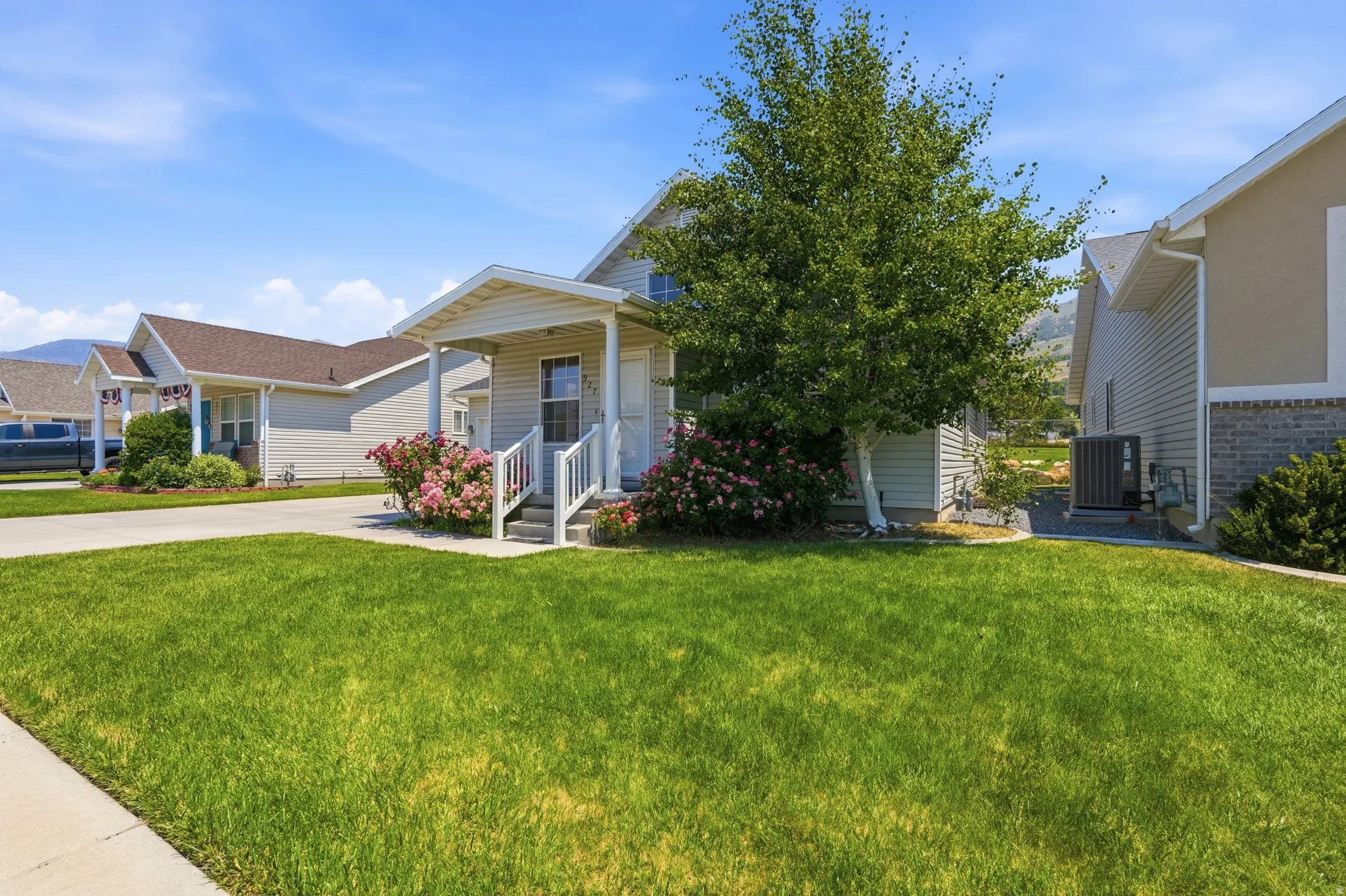 Front exterior featuring siding, an inviting covered porch, lush green lawn, mature tree, and vibrant shrubbery