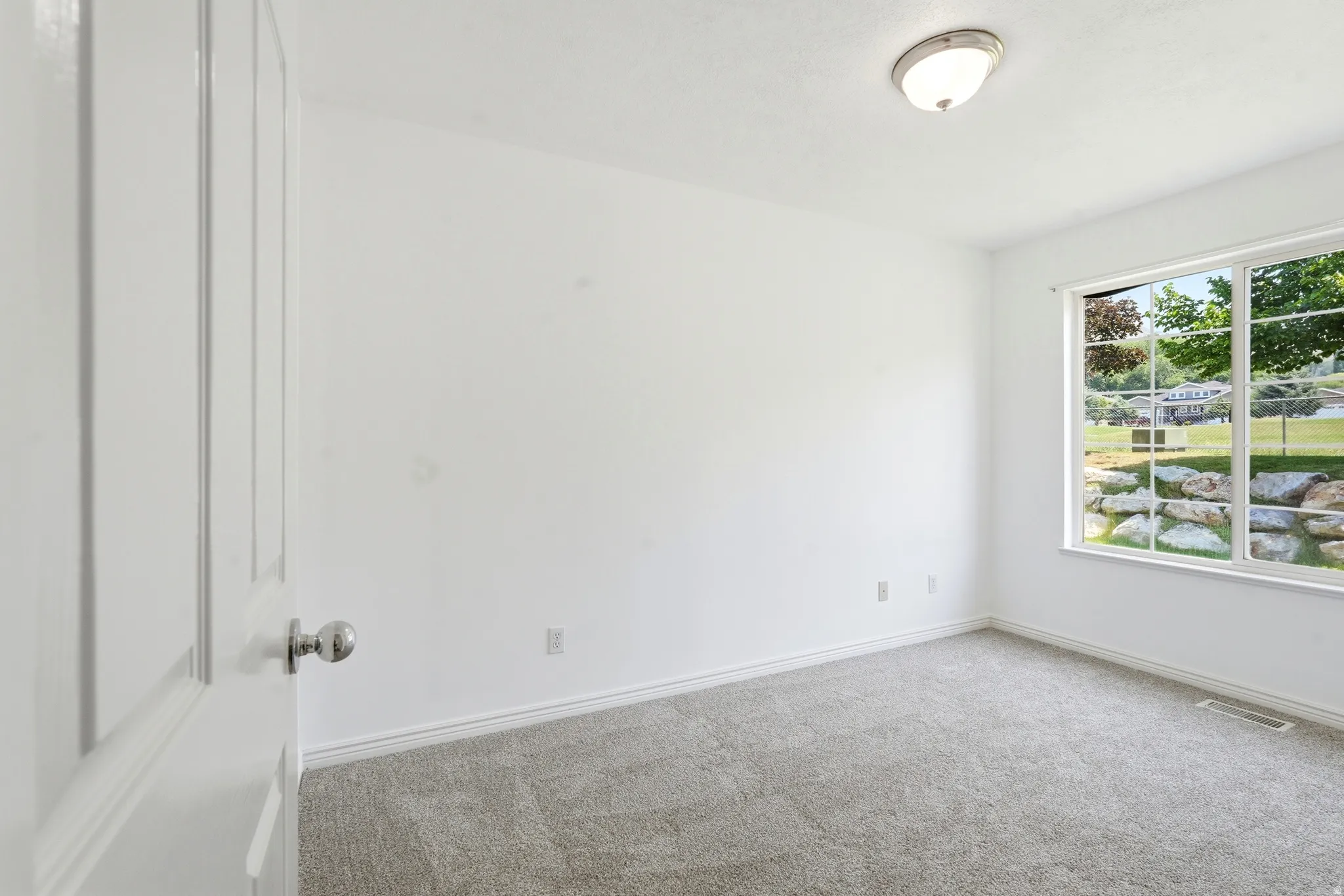 Bright room featuring light gray carpeting, white walls, and a multi-pane window overlooking a landscaped exterior with trees and rock elements