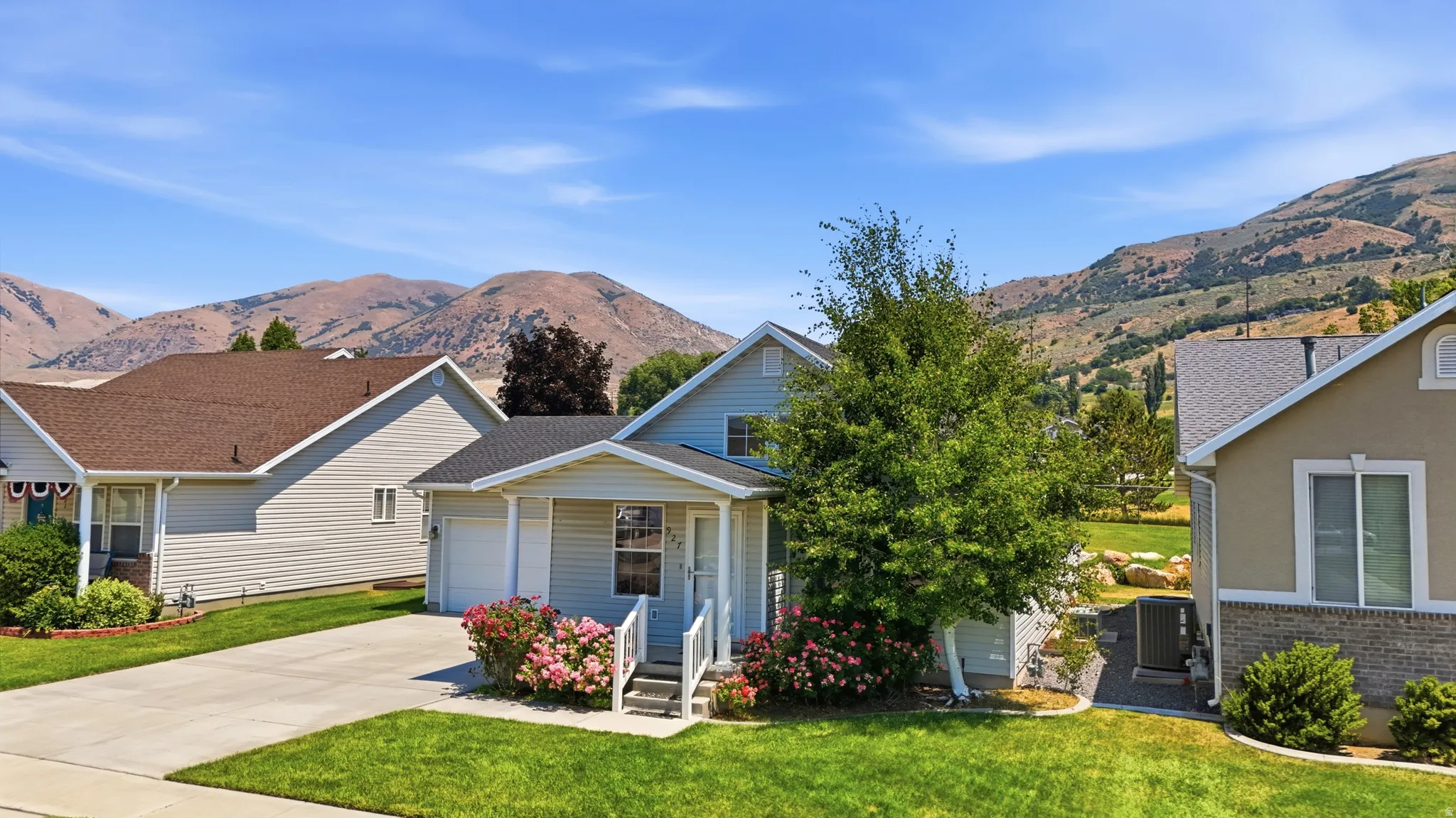 Light blue siding residence with a single-car garage and white trim, featuring a front lawn with mature trees and flowering shrubs