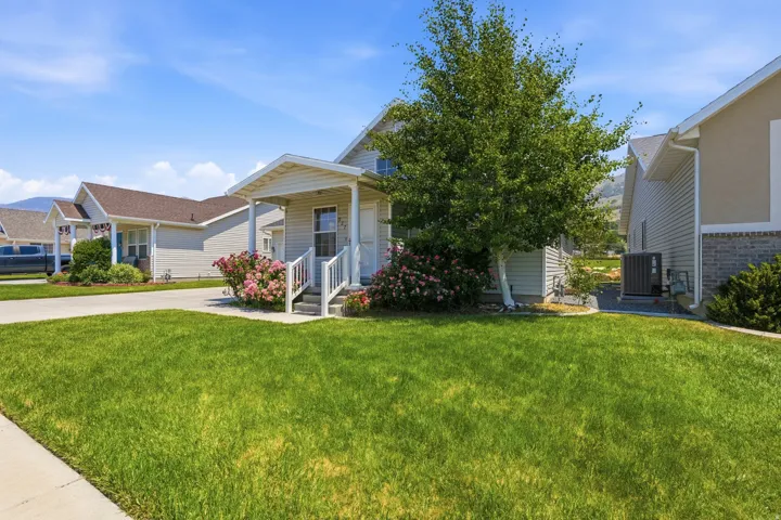 Front exterior featuring siding, an inviting covered porch, lush green lawn, mature tree, and vibrant shrubbery