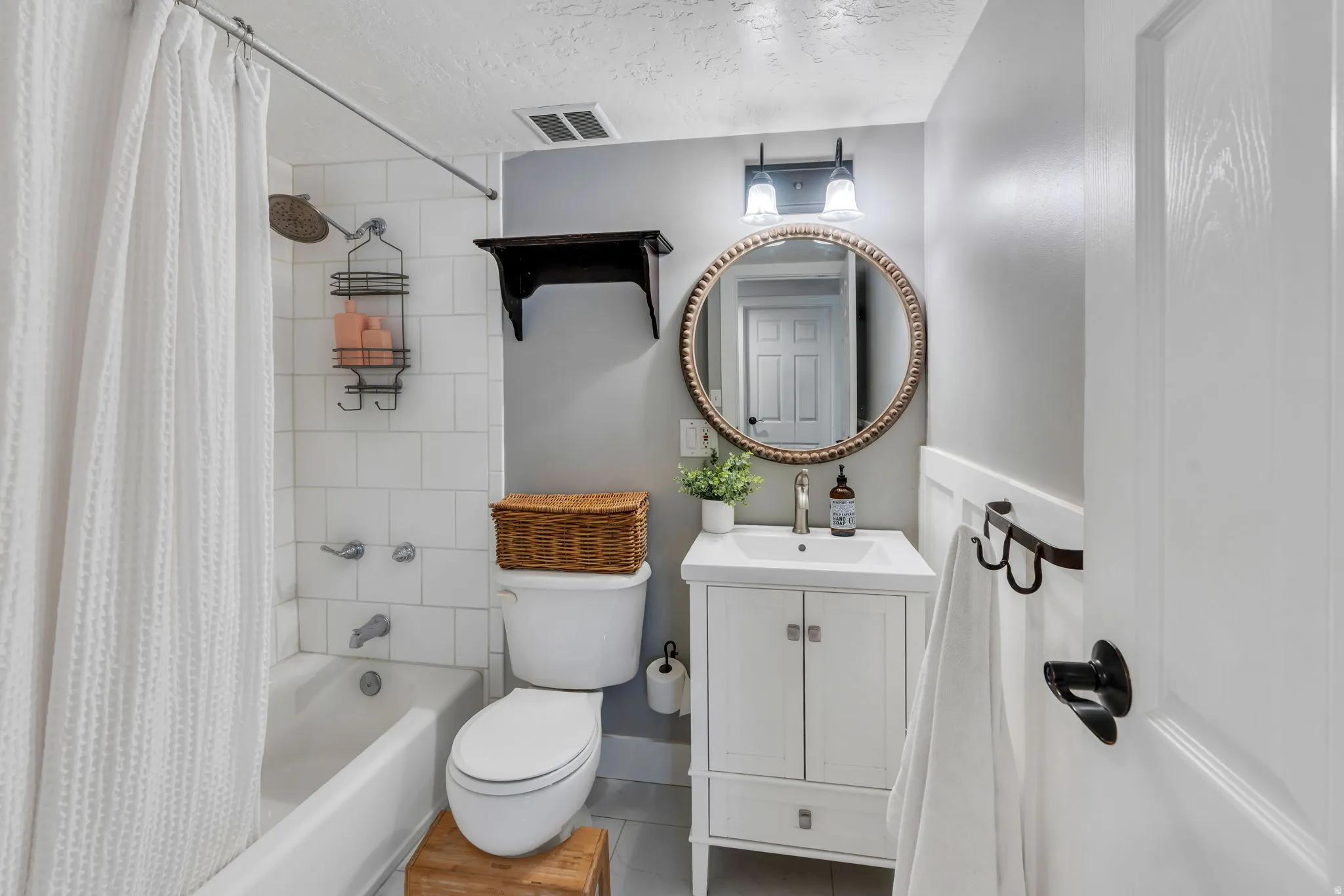 Main level full bathroom featuring a white subway tile tub surround, a white vanity with integrated sink, a round decorative mirror, and a two-bulb wall-mounted light fixture