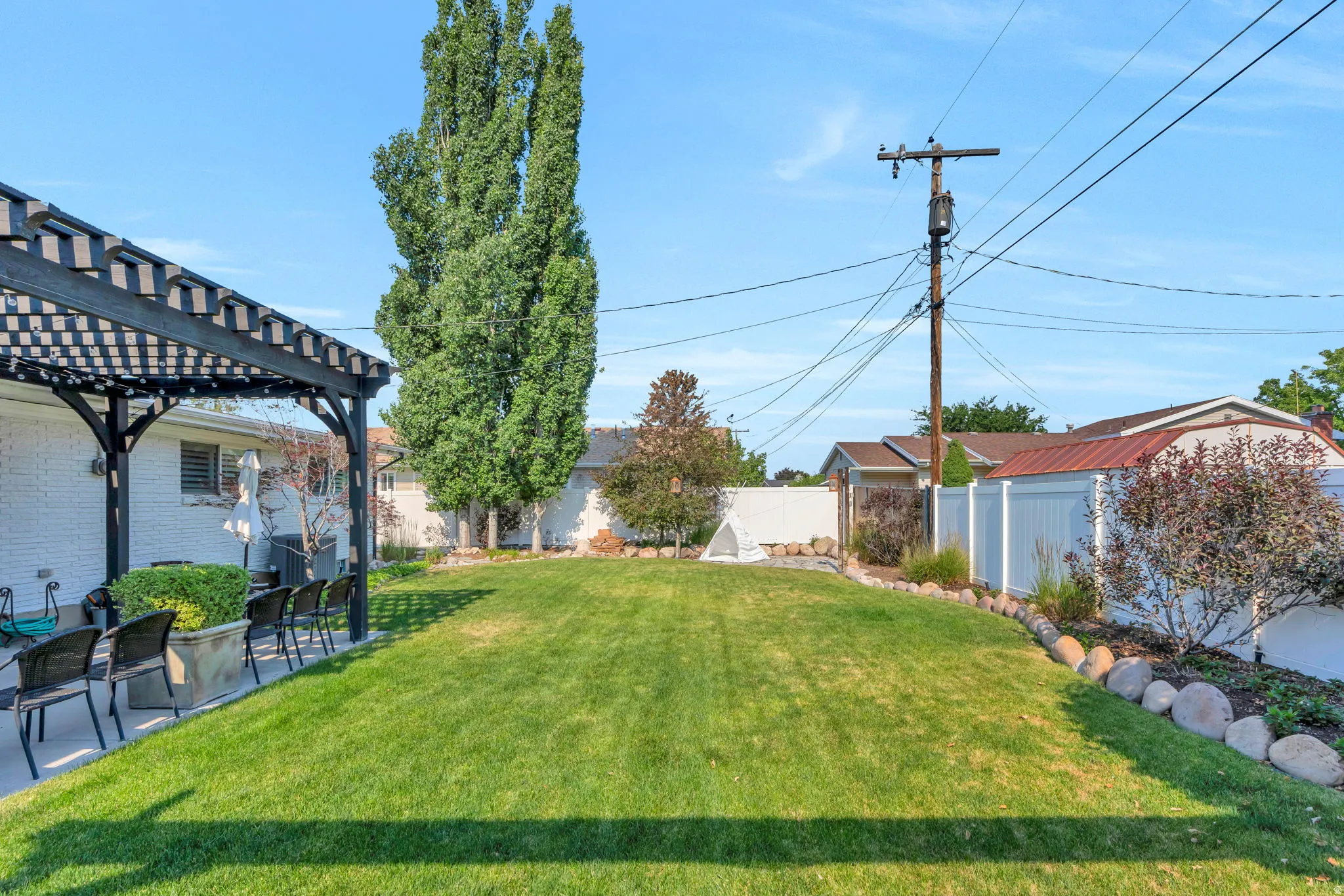 Expansive backyard featuring a large green lawn, a covered patio with a black pergola, white brick exterior, and a white privacy fence