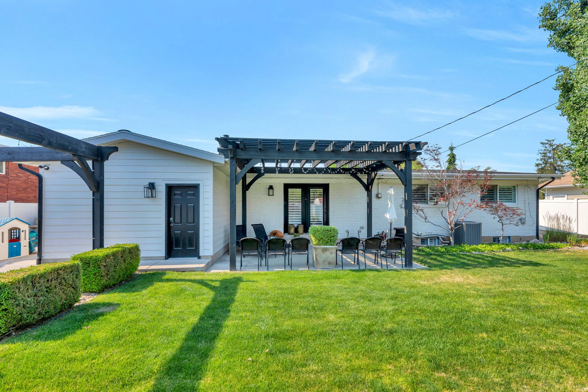 Spacious backyard featuring a manicured lawn, a large patio with a black pergola, and white brick exterior