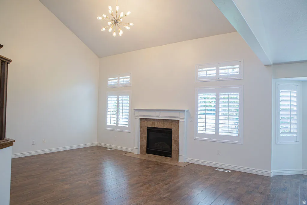 Vaulted ceiling featuring a contemporary chandelier, a fireplace with a stone surround and white mantel, multiple windows with white plantation shutters, and wood-finish flooring