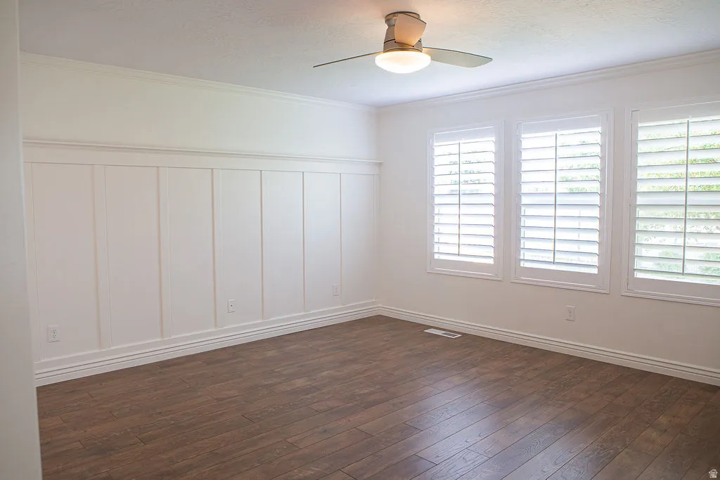 Room featuring wood-finish flooring, white board and batten wainscoting, crown molding, three windows with plantation shutters, and a ceiling fan with integrated lighting