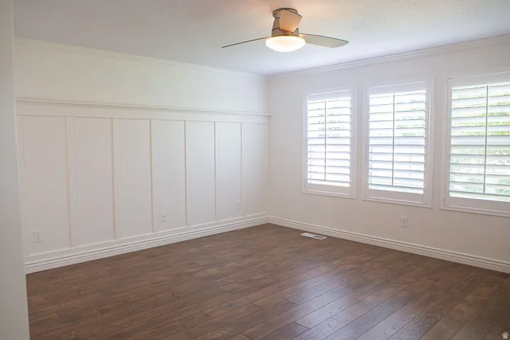 Room featuring wood-finish flooring, white board and batten wainscoting, crown molding, three windows with plantation shutters, and a ceiling fan with integrated lighting
