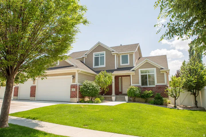 Two-story residence featuring a combination of brick and siding, a multi-car garage with white doors, a concrete driveway, and a manicured lawn with mature trees and shrubbery