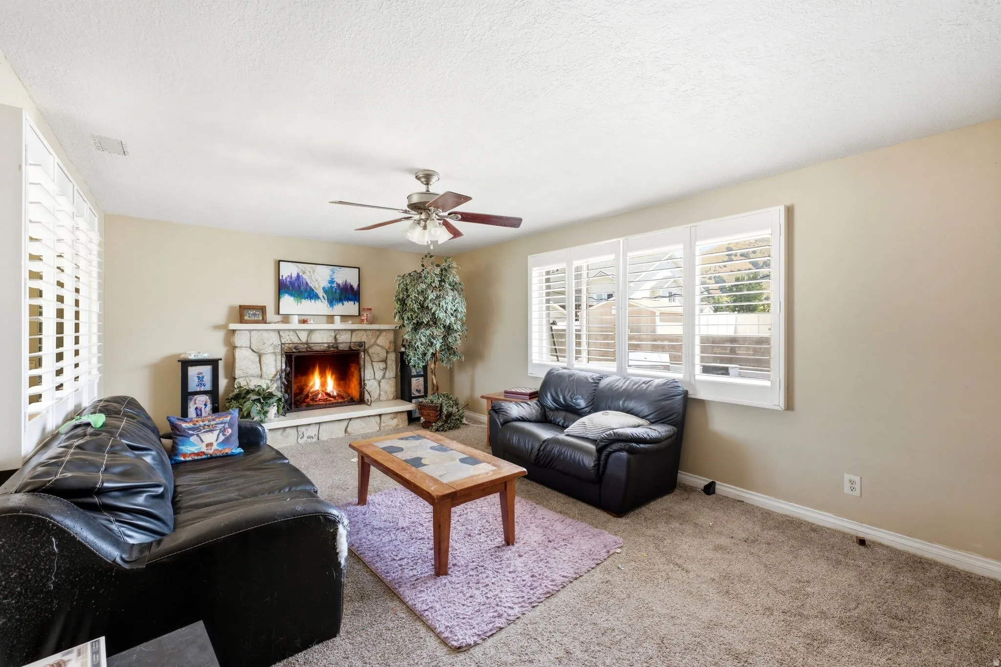 Living area featuring a stone-clad fireplace with a hearth, multiple windows with white plantation shutters, a ceiling fan with integrated lighting, and neutral-toned wall paint