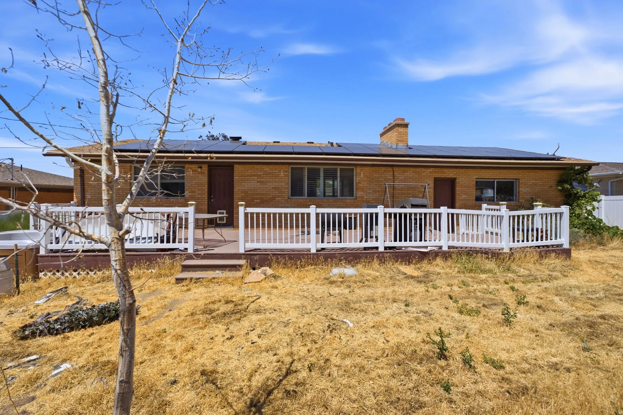 Rear exterior featuring a brick facade, full-length elevated deck with white railing, integrated solar panels, and a prominent brick chimney