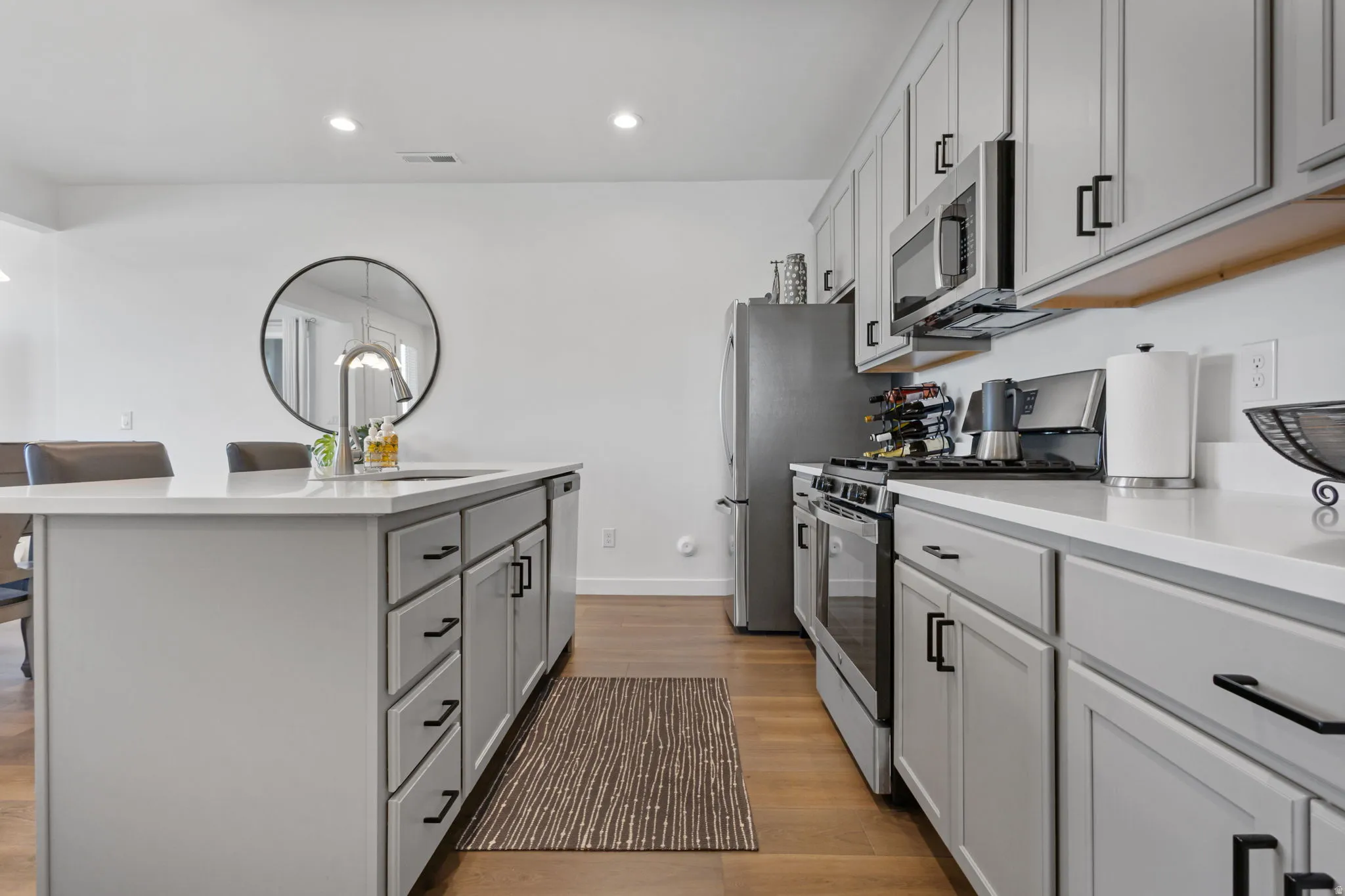 Kitchen featuring light gray cabinetry, stainless steel appliances, wood-finish flooring, white countertops, and a central island with an undermount sink