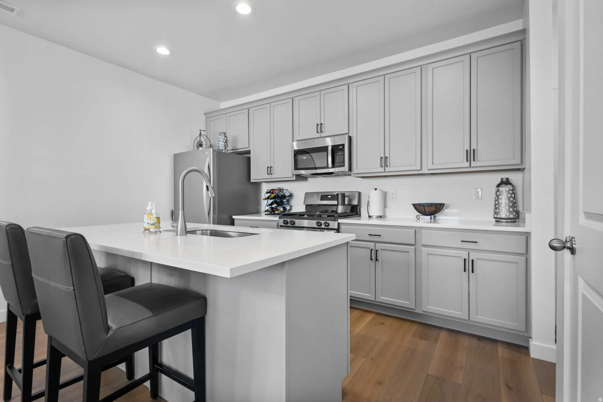 Kitchen featuring a central island with a sink, light gray cabinetry, white countertops, stainless steel appliances, and wood-finish flooring
