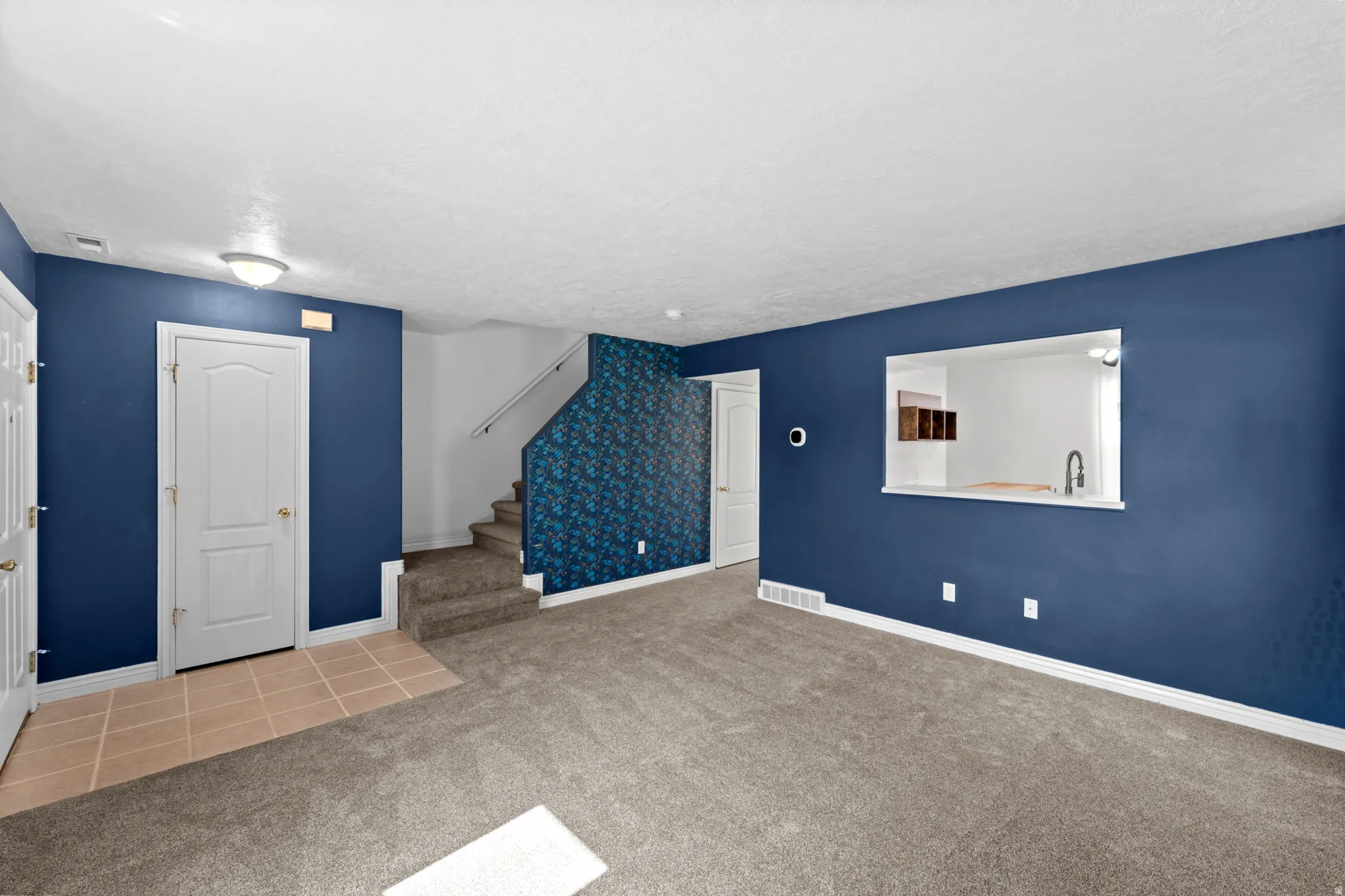 Spacious living area featuring carpet flooring, a pass-through window to the kitchen, and a staircase with carpeted treads and a white handrail