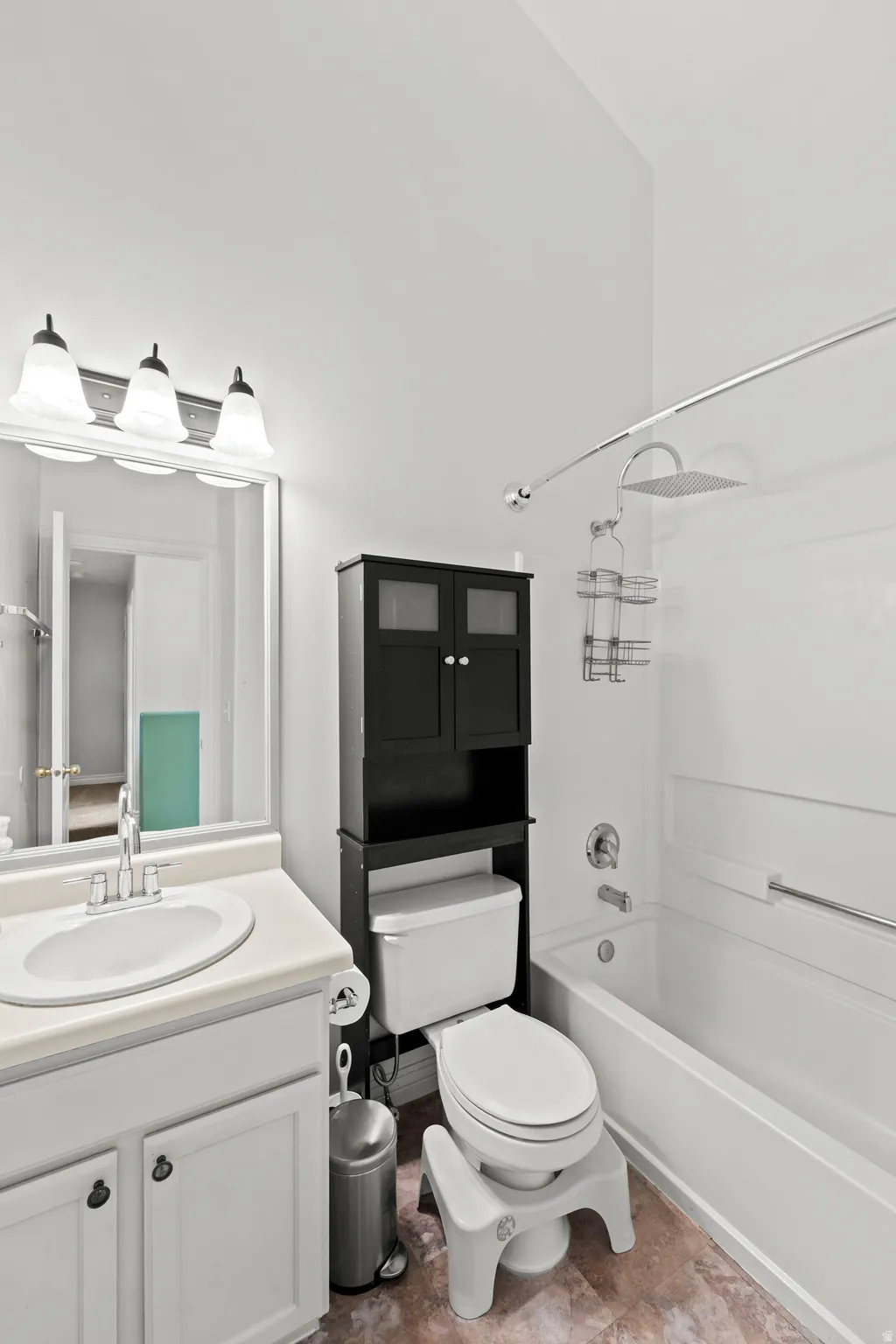 Bathroom featuring a white vanity with an oval sink, a wall-mounted light fixture with three sconces, a combination shower and bathtub, a rain shower head, and a black over-the-toilet storage cabinet