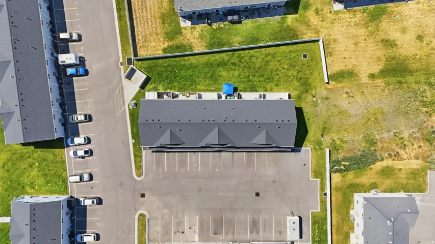 Multi-unit residential structure featuring a dark shingle roof, integrated parking lot with marked spaces, surrounding green spaces, paved driveways, and a nearby row of similar buildings