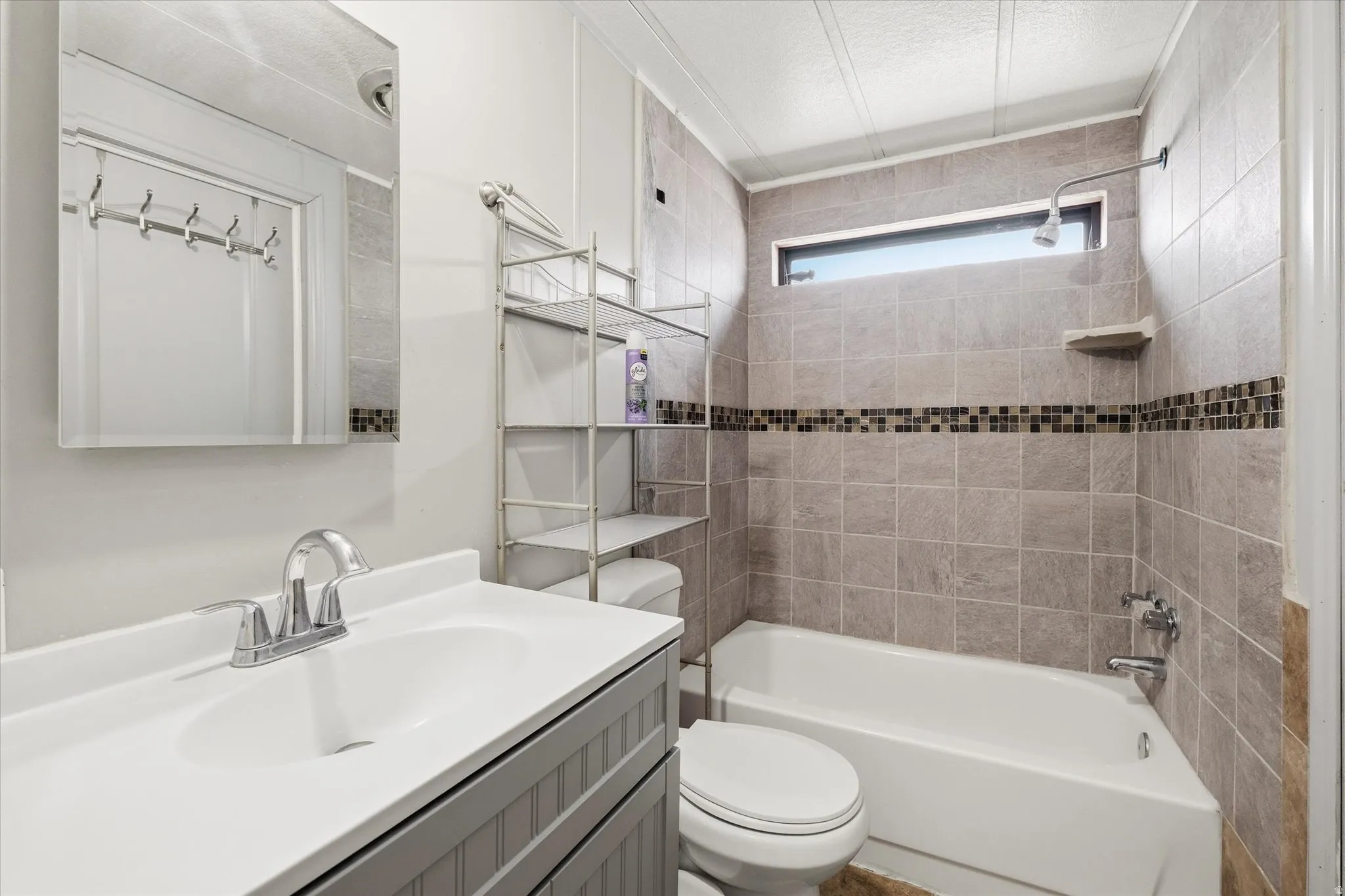Bathroom featuring a single vanity with a white countertop and chrome faucet, a rectangular mirror, and a tiled shower/tub combination with a mosaic accent band