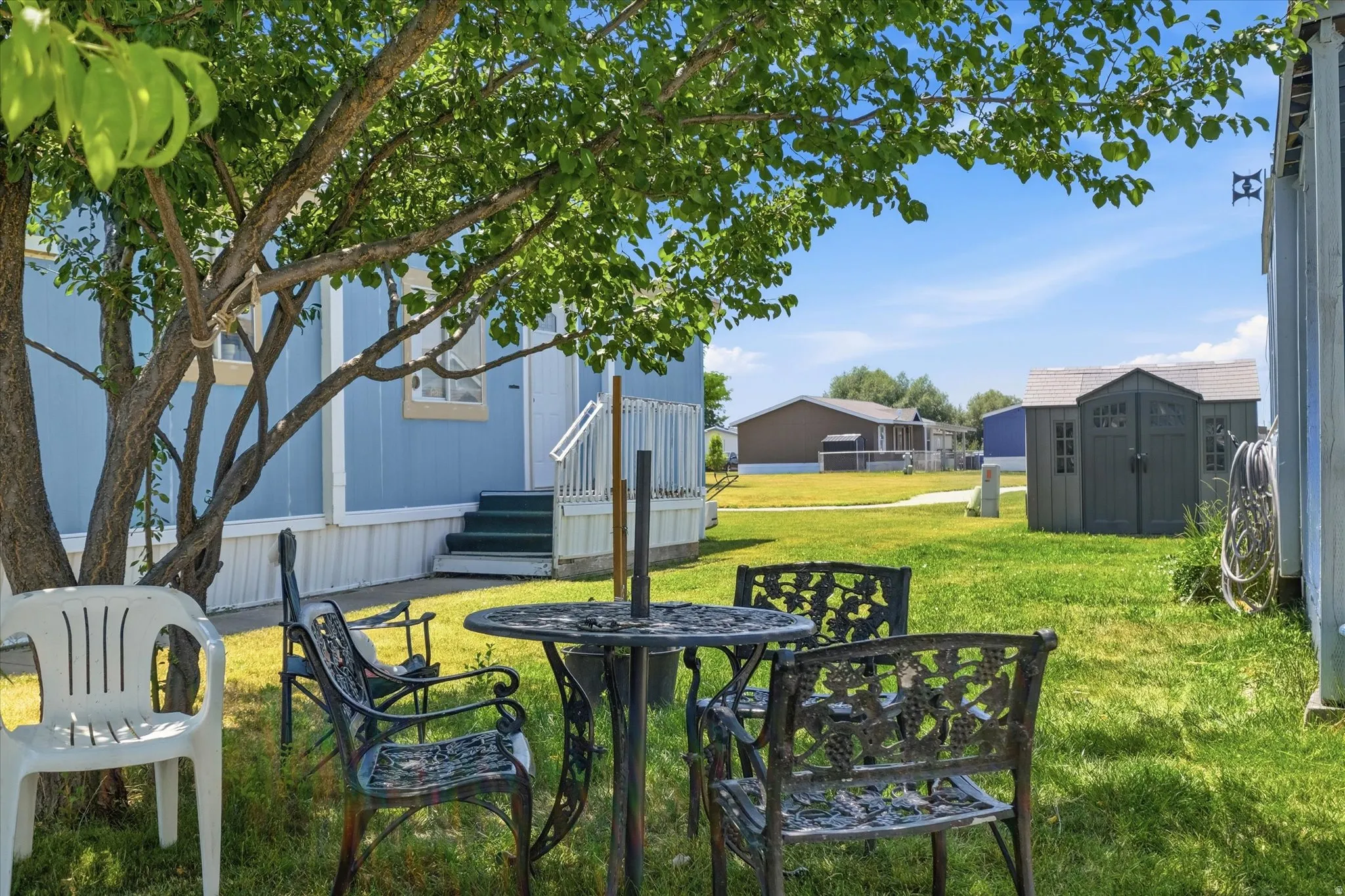 Expansive grassy yard featuring a mature tree, a light blue exterior structure with white trim, a storage shed, and additional structures in the background