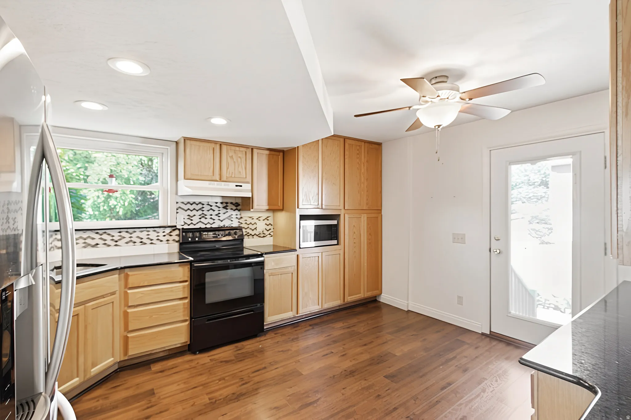Kitchen featuring wood-finish flooring, light wood cabinetry, black countertops, and a mosaic tile backsplash