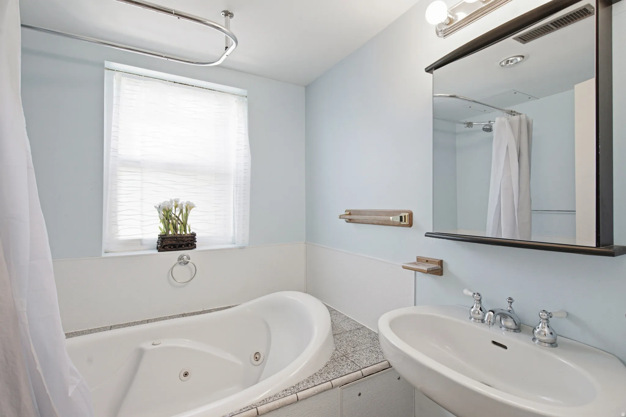 Bathroom featuring a jetted tub, a window with blinds, a wall-mounted sink with chrome fixtures, a framed mirror, and a wall-mounted light fixture