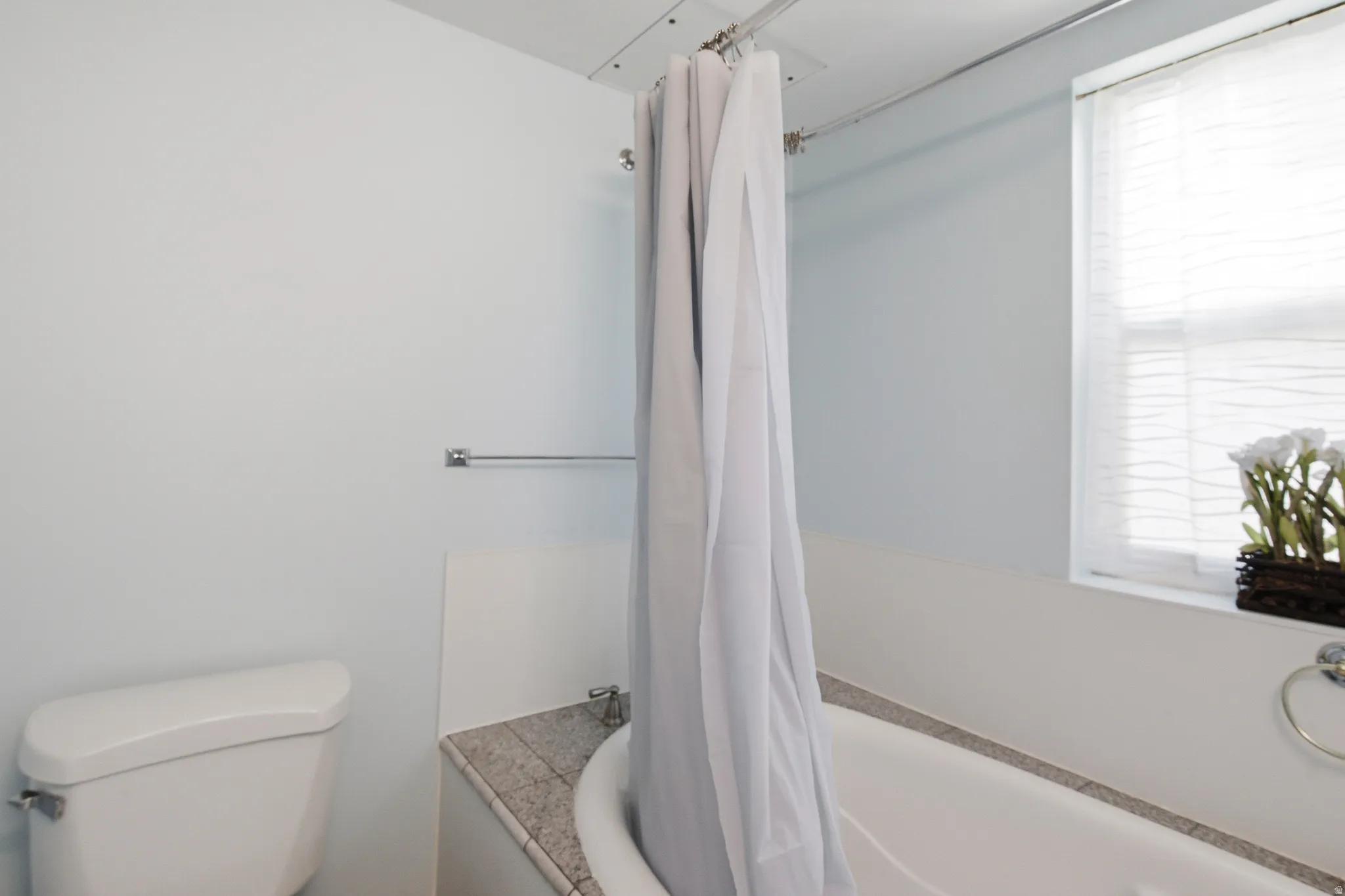 Bathroom featuring a white porcelain toilet, a white bathtub with a stone-finish surround, a window with horizontal blinds, and a chrome towel ring