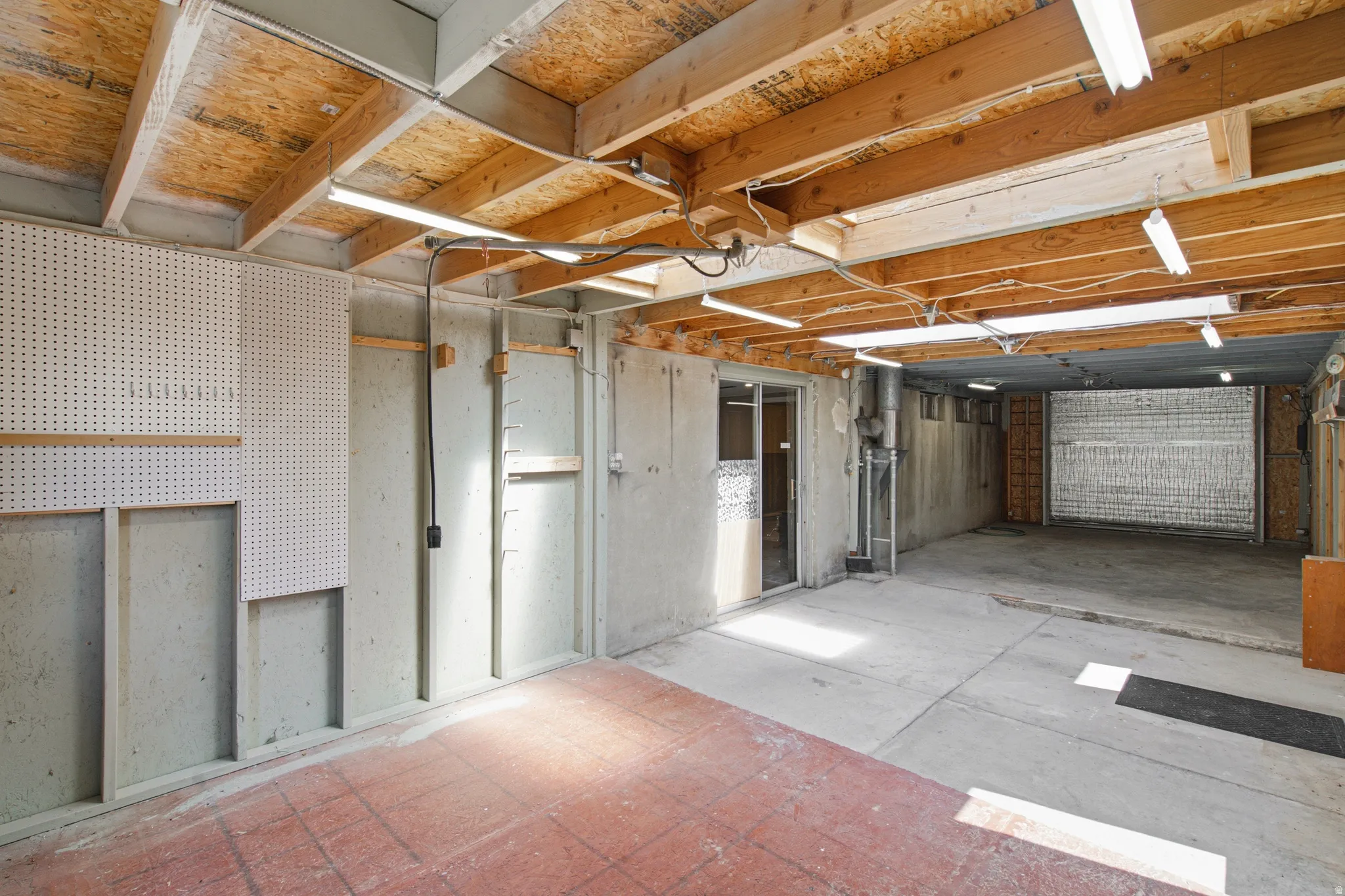 Utility space featuring exposed wood ceiling joists, concrete flooring, fluorescent lighting, a sliding glass door, and pegboard wall storage