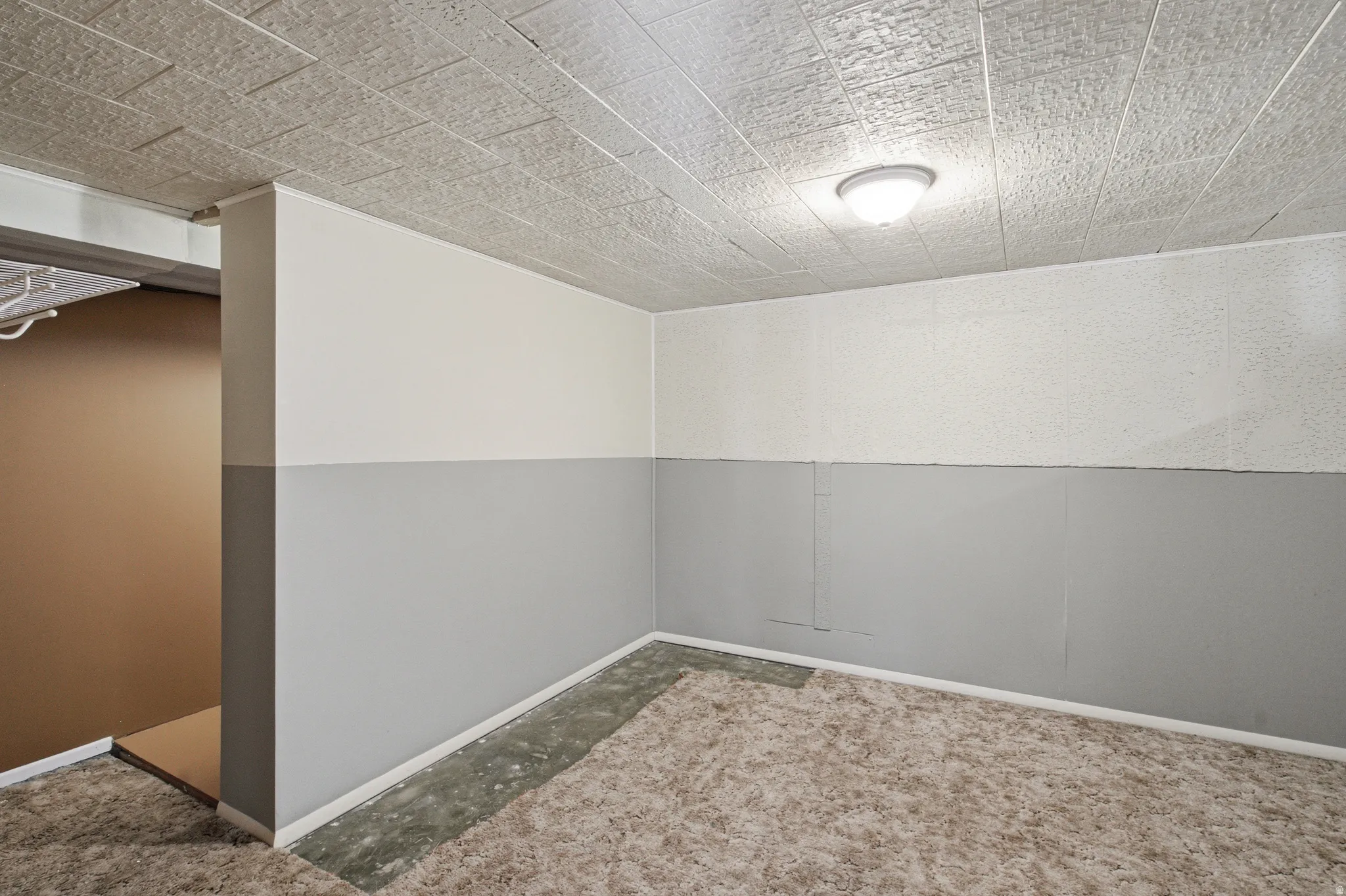 Interior space featuring a textured acoustic tile ceiling, overhead flush-mount lighting, two-tone painted walls with a neutral upper portion and grey lower paneling, and a neutral patterned carpet partially covering the floor