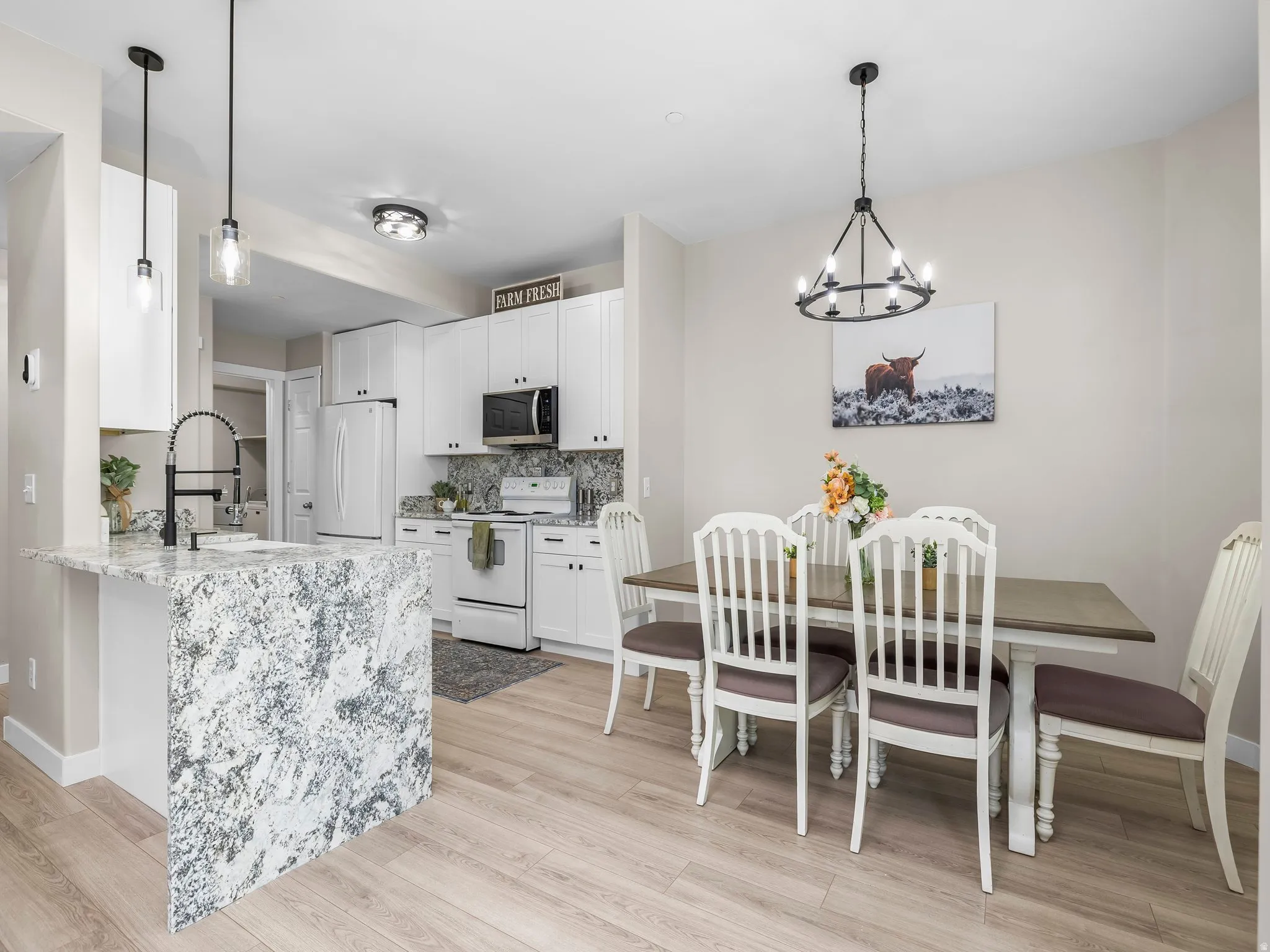Open-concept kitchen and dining area featuring light wood-finish flooring, white cabinetry, a kitchen island with a stone countertop and waterfall edge, and a dark-finish chandelier