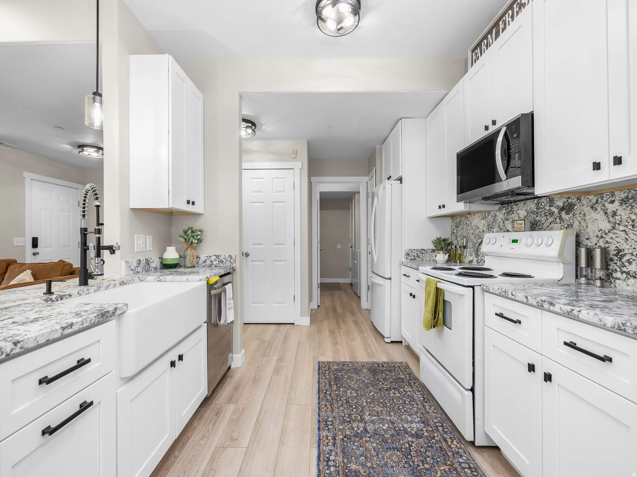Kitchen featuring white shaker-style cabinetry, light-toned wood-finish flooring, a white farmhouse sink with a black pull-down faucet, stainless steel appliances, and mottled stone countertops with a matching backsplash