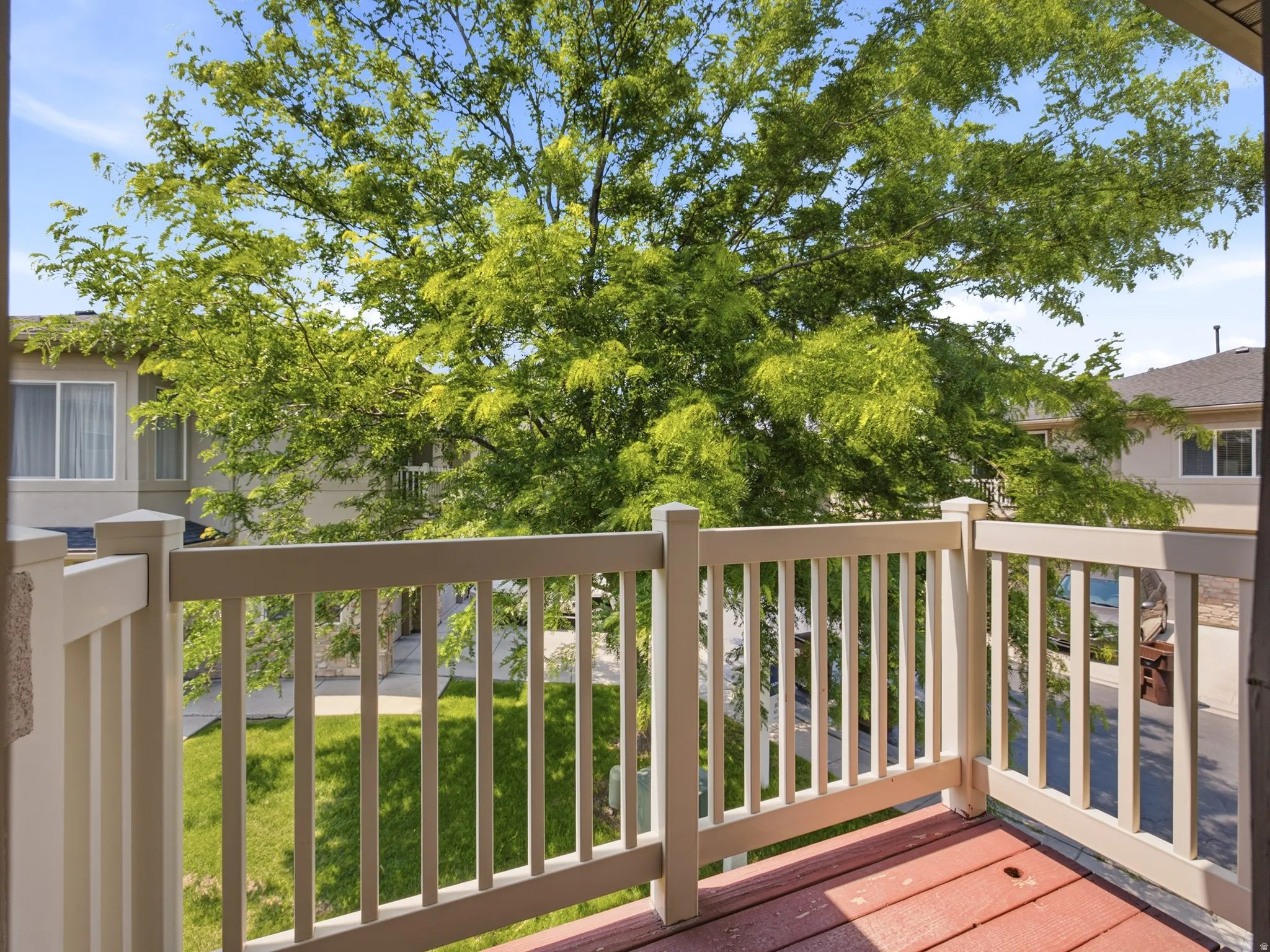 Outdoor balcony with a red-toned floor and light-colored railing, overlooking a green space with mature trees