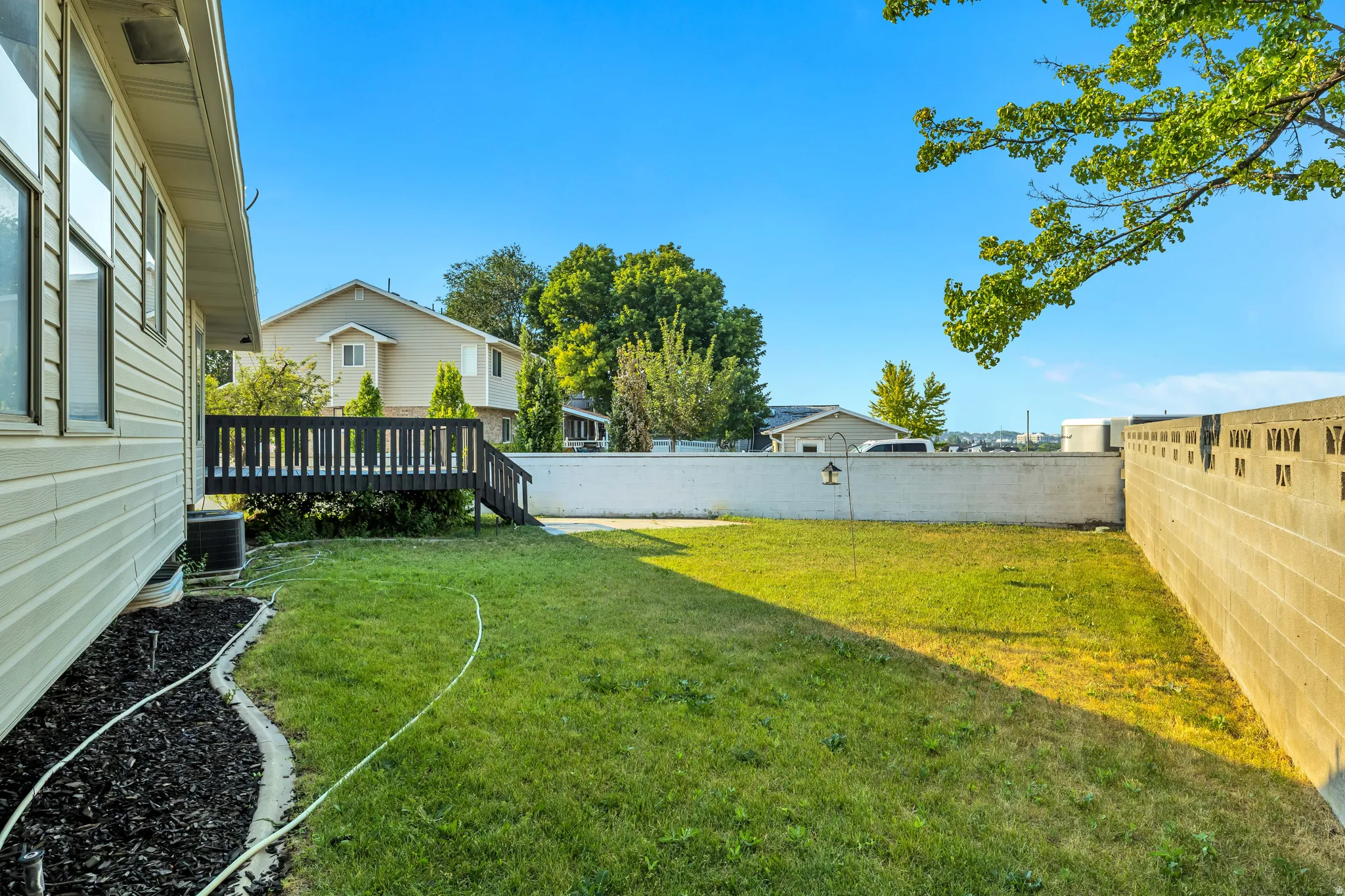 Expansive grassy yard area featuring a private wood-stained deck with railing, a decorative landscape bed with dark mulch, and perimeter fencing