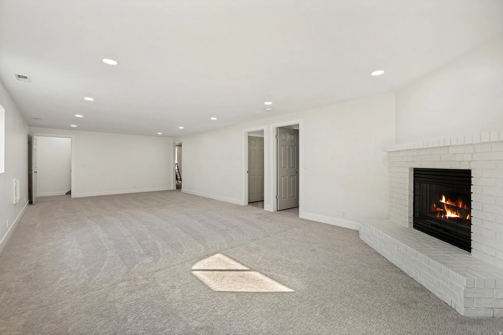 Spacious carpeted room featuring a white brick fireplace with a raised hearth, recessed lighting, white walls, and multiple interior doorways
